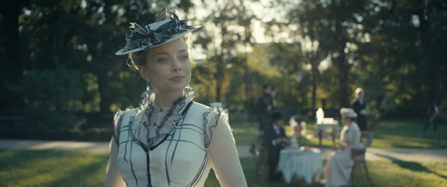 kobieta w eleganckiej sukni czarno-białej, w kapeluszu, z delikatnym uśmiechem, na tle drzew i ustawionych pod nimi stolików.